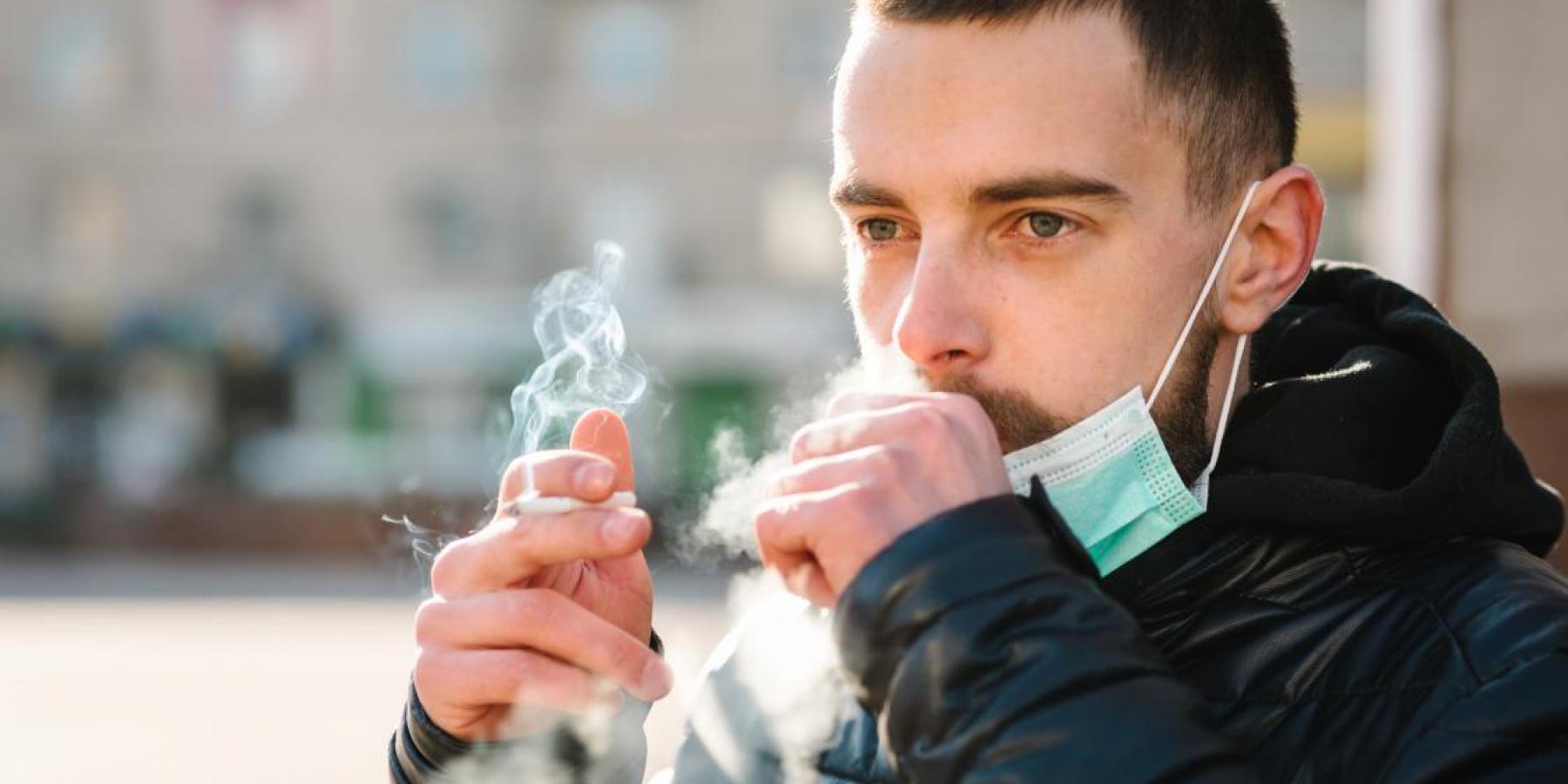 Closeup man with mask during COVID-19 pandemic coughing and smoking a cigarette at the street